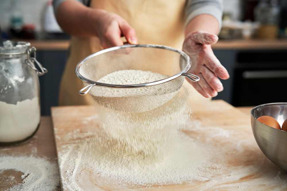 A close up of a person sifting flour over a cutting board in a kitchen