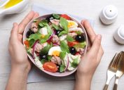 Woman holding bowl of delicious salad with canned tuna at white wooden table, top view