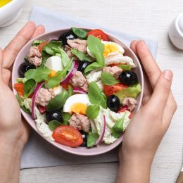 Woman holding bowl of delicious salad with canned tuna at white wooden table, top view