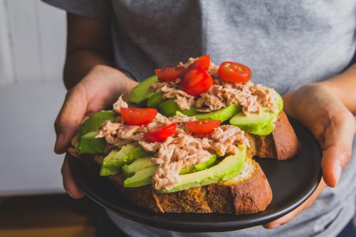 Person holding a sandwich with tuna fish and sliced avocado on top tomato