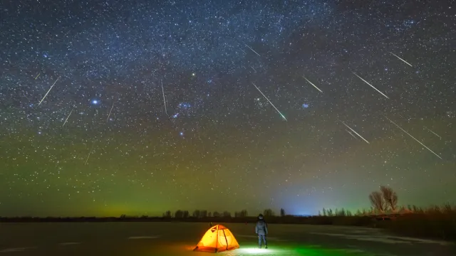 A person standing outside their tent looking up at shooting starts during a meteor shower