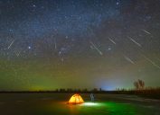 A person standing outside their tent looking up at shooting starts during a meteor shower