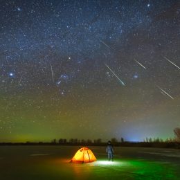 A person standing outside their tent looking up at shooting starts during a meteor shower