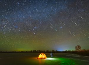 A person standing outside their tent looking up at shooting starts during a meteor shower