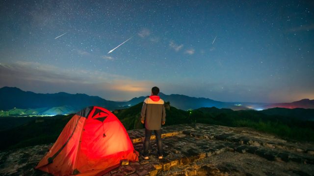 A person watching a meteor shower in the night sky standing next to their tent