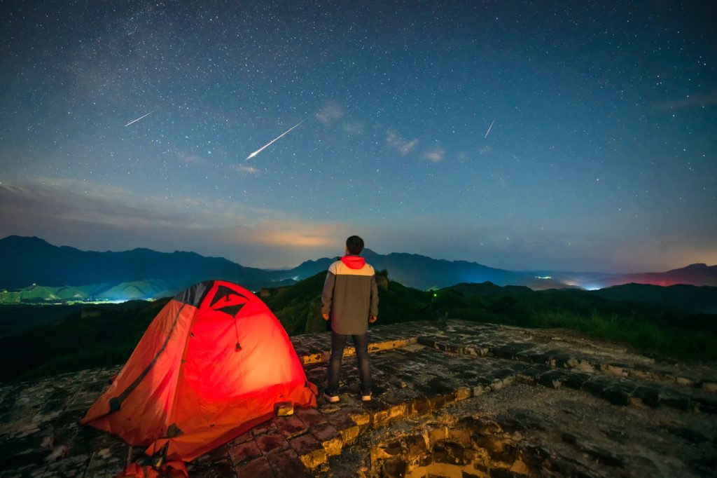 A person watching a meteor shower in the night sky standing next to their tent