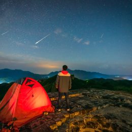 A person watching a meteor shower in the night sky standing next to their tent