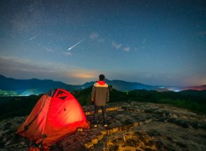 A person watching a meteor shower in the night sky standing next to their tent