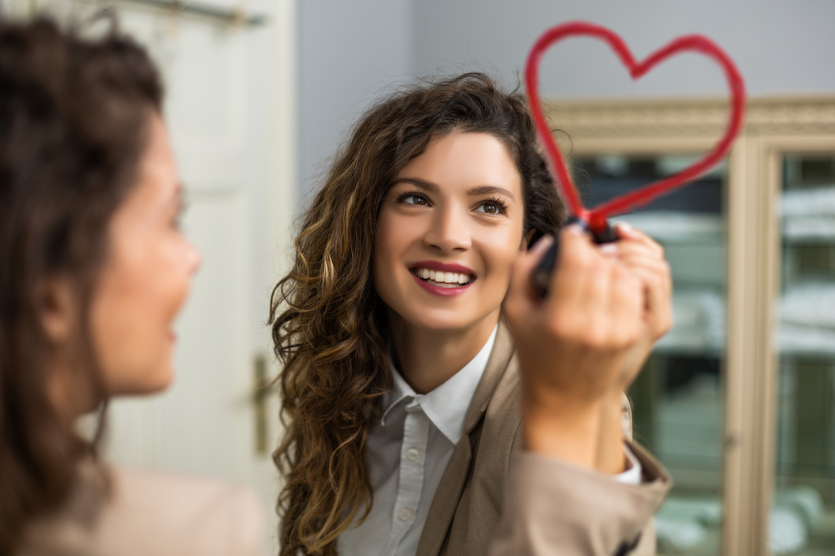 Smiling businesswoman is drawing heart with lipstick on the mirror while preparing for work.