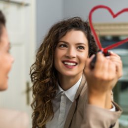 Smiling businesswoman is drawing heart with lipstick on the mirror while preparing for work.
