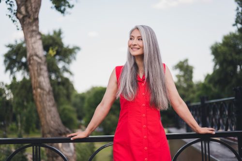 A smiling woman with long gray hair wearing a red dress leans against a railing outside