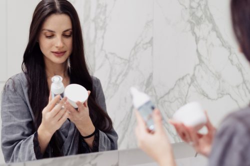 Attractive young woman with perfect long hair looking at bottle of face cleansing gel and jar with moisturizing cream during her morning routine at bathroom.