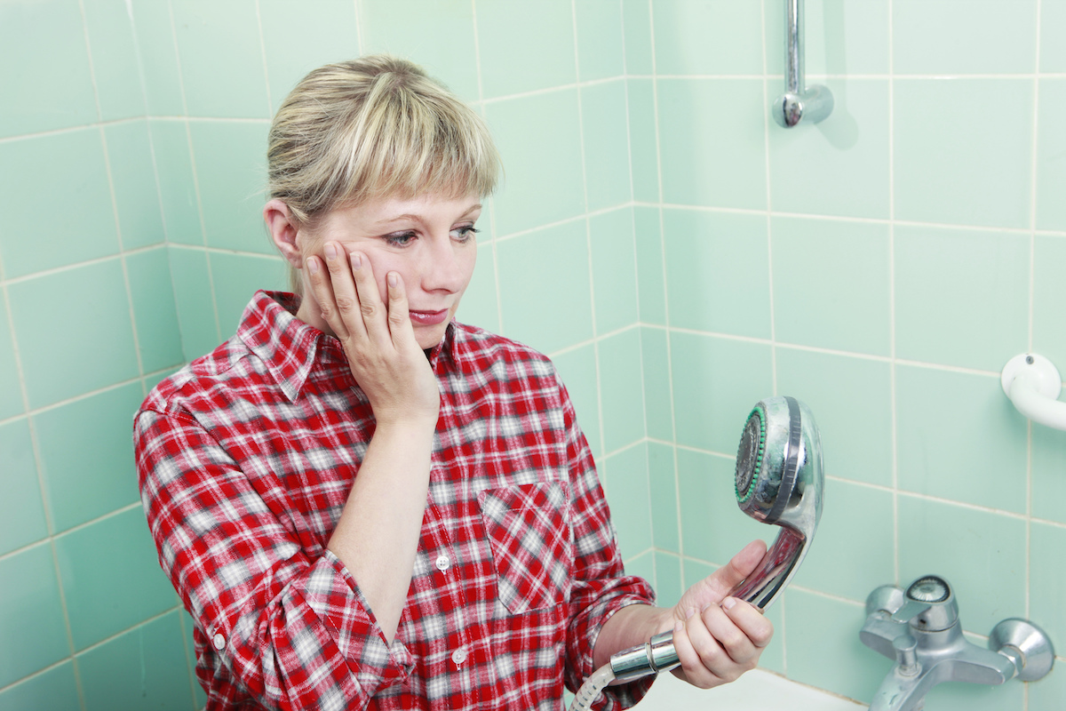 Woman in a red plaid shirt standing in her seafoam green-tiled shower, looking down at her dirty shower head.