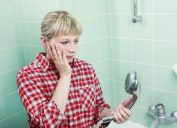 Woman in a red plaid shirt standing in her seafoam green-tiled shower, looking down at her dirty shower head.