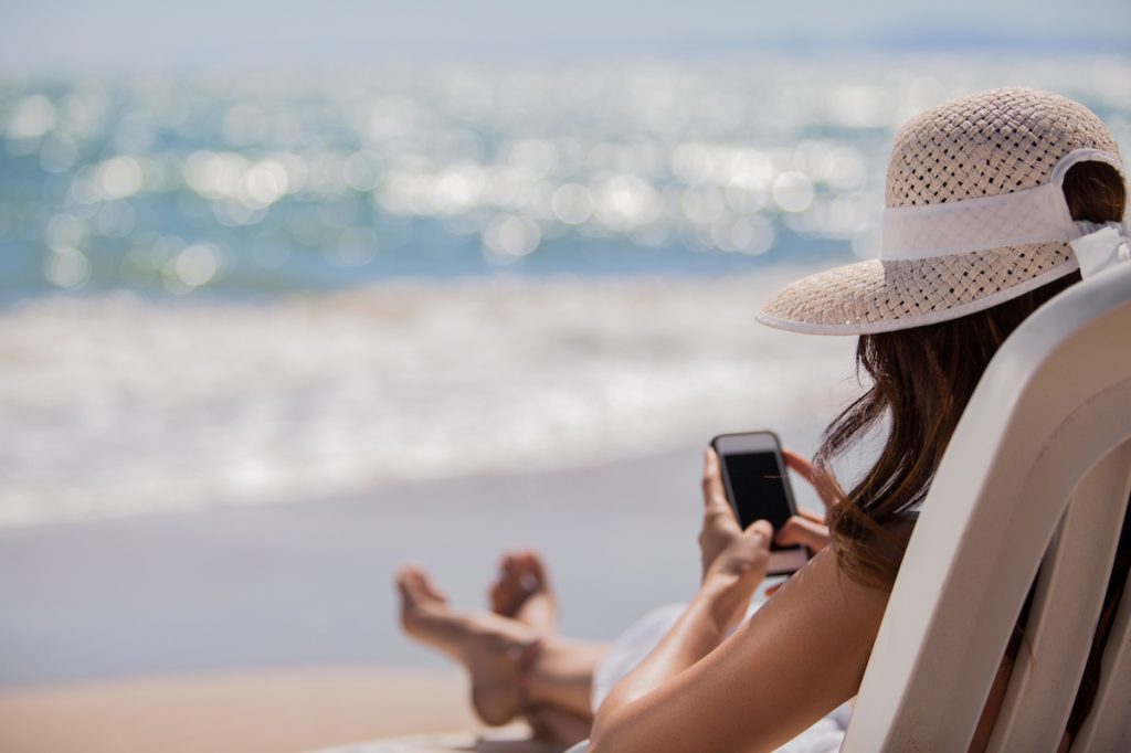 A young woman wearing a hat uses her smartphone by the wavebreak of a beach on a sunny day.