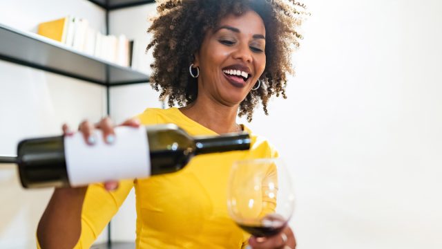 A smiling woman wearing a bright yellow shirt pours herself a glass of red wine.