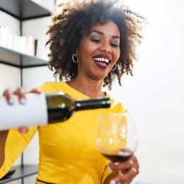 A smiling woman wearing a bright yellow shirt pours herself a glass of red wine.