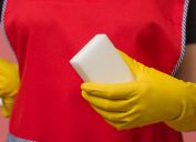 white melamine sponge in the hand of a cleaning woman in protective gloves and red apron, close-up