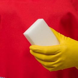 white melamine sponge in the hand of a cleaning woman in protective gloves and red apron, close-up