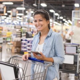 A woman smiling while pushing her cart through a warehouse bulk goods store and holding her smartphone