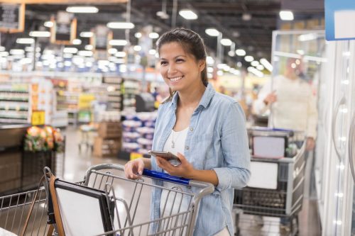 A woman smiling while pushing her cart through a warehouse bulk goods store and holding her smartphone