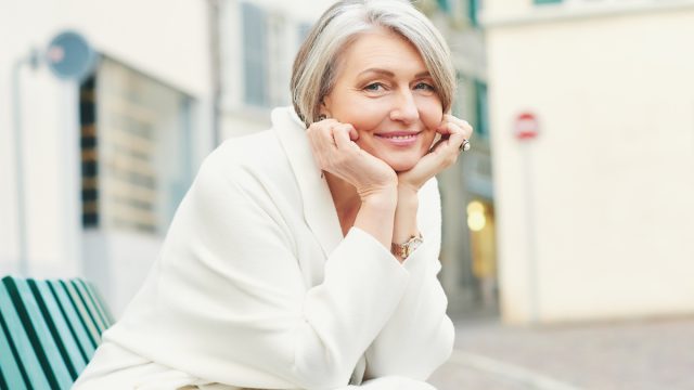 50-60 year old woman, wearing a white jacket, sitting on a bench outside