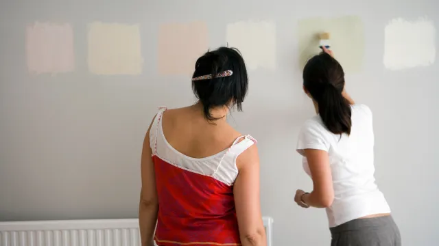 Rear view of two women testing different paint shades over the wall.