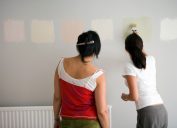 Rear view of two women testing different paint shades over the wall.