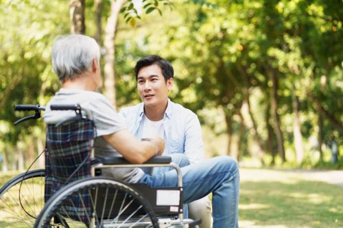 young asian adult son chatting with wheel chair bound father outdoors in park
