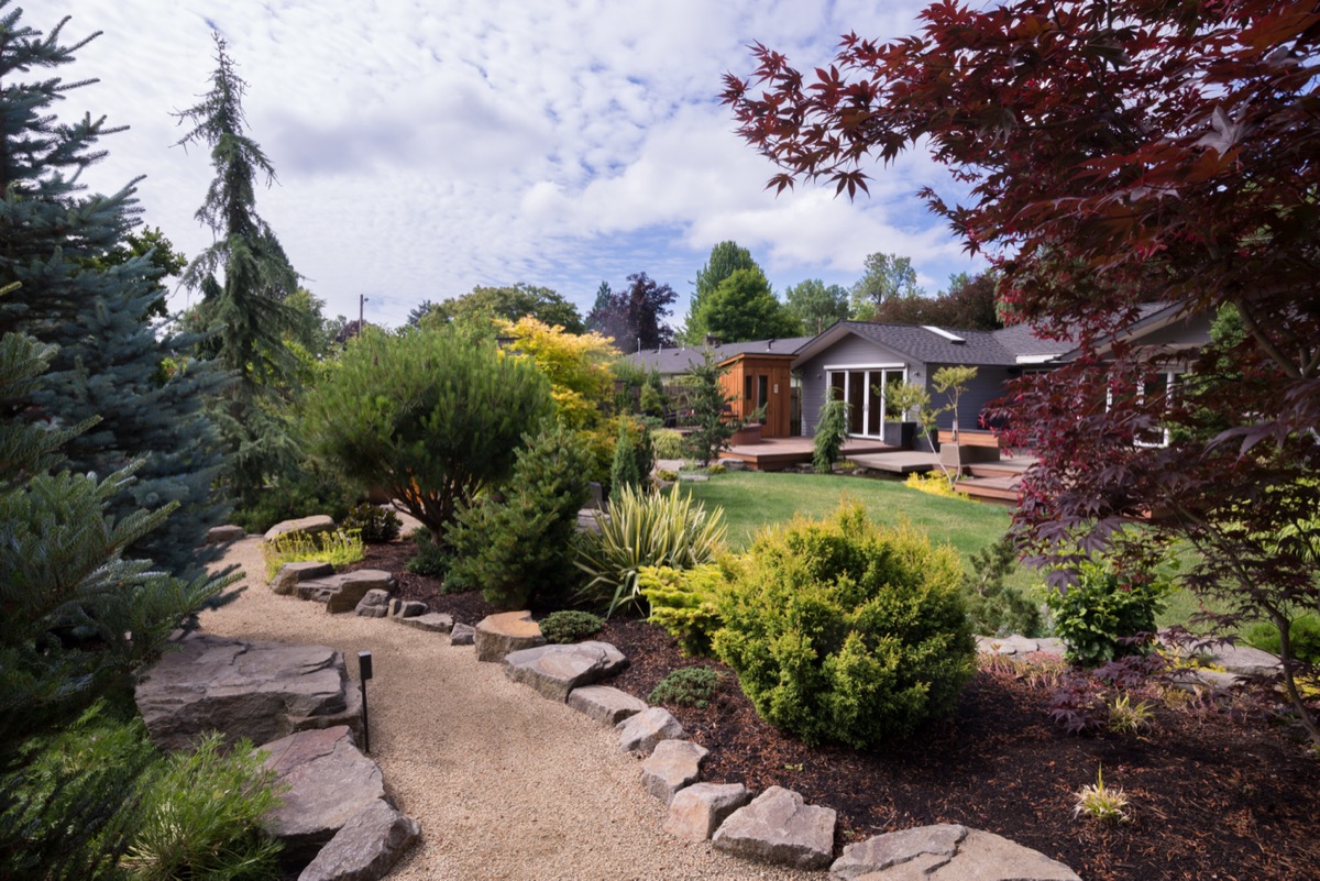 A path meanders between naturally sculptured small rocks, with a variety of trees and shrubs on either side. In the background, a beautiful contemporary home can be seen beyond a lush green lawn.