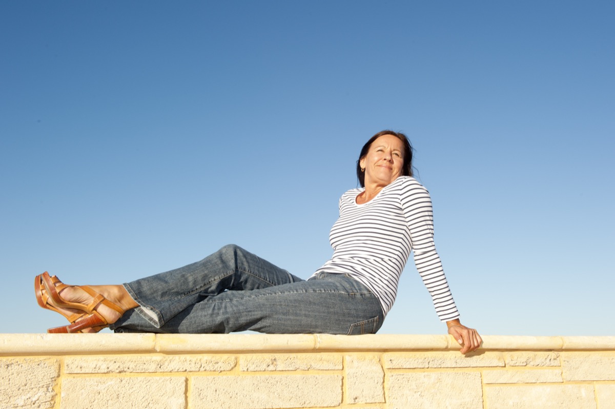 Portrait pretty mature woman sitting on a sunny day relaxed and happy on limestone wall, isolated with clear blue sky as background and copy space.