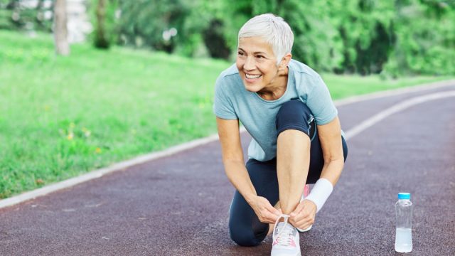 Portrait of a happy active beautiful senior woman posing fixing shoelace on her running shoe after exercising outdoors