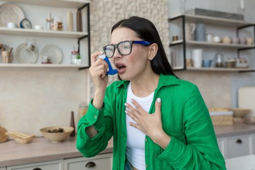 Sick young woman at home with asthma, using inhaler to ease breathing and treatment, brunette in kitchen wearing glasses and green shirt