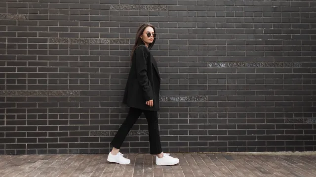 Trendy young woman in stylish jacket in vintage jeans in white sneakers in sunglasses walks near brick wall in city. Attractive girl in casual black clothes is walking on street. American style.