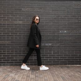 Trendy young woman in stylish jacket in vintage jeans in white sneakers in sunglasses walks near brick wall in city. Attractive girl in casual black clothes is walking on street. American style.