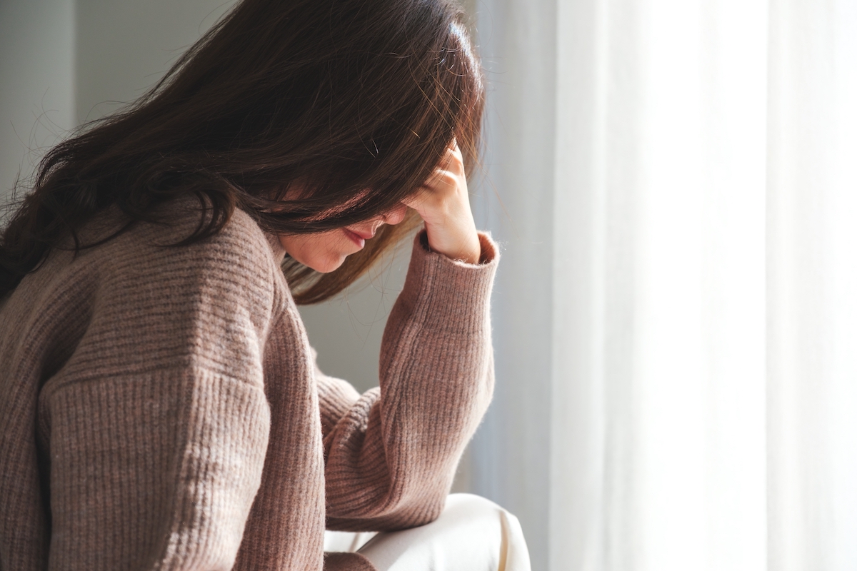 Closeup of a woman wearing a beige sweater sitting with her head in her hand and her long brown hair covering her face