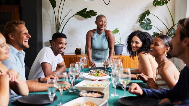 Young woman laughing at the head of a table while hosting a dinner party for a group of diverse young friends at her home