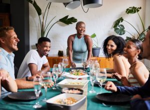 Young woman laughing at the head of a table while hosting a dinner party for a group of diverse young friends at her home