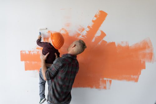 Father holds his son up so he can paint the wall orange.