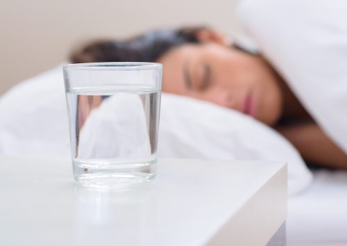 Một cốc nước trên bàn cạnh giường ngủ với một người phụ nữ đang ngủ ở phía sau