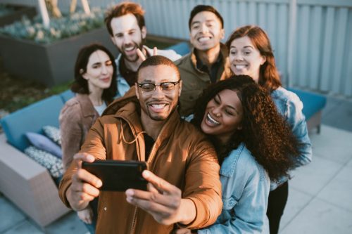A group of friends have a party or celebration outside in downtown Los Angeles, California. One of them takes a selfie of the group on their smartphone to share on social media.