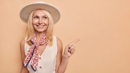 A happy middle-aged blonde woman wearing a beige dress, printed scarf, and beige sun hat.