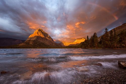 Fire blazes across the sky and a rainbow appears as a late season storm powers its way onto Swiftcurrent Lake raising the water into froth and wave in one of the most beautful places on earth, Glacier National Park located in Montana.
