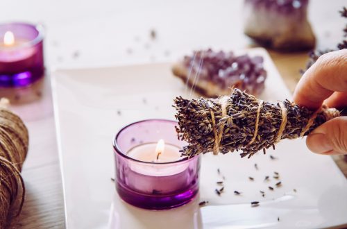 Person holding homemade herbal lavender (lavendula) smudge stick with smoke coming out, candles and amethyst crystal clusters for decoration