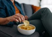 Young unrecognizable man at home, sitting on sofa and taking a potato chip from a bowl placed on a sofa
