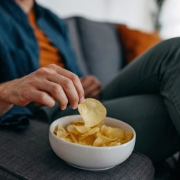 Young unrecognizable man at home, sitting on sofa and taking a potato chip from a bowl placed on a sofa