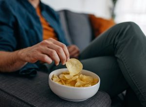 Young unrecognizable man at home, sitting on sofa and taking a potato chip from a bowl placed on a sofa