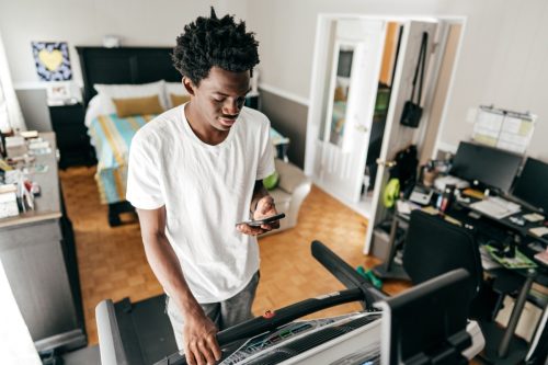 Một thanh niên sử dụng máy chạy bộ trong phòng ngủ của mình, anh ấy cũng kết nối ứng dụng điện thoại di động của mình với cài đặt chạy bộ