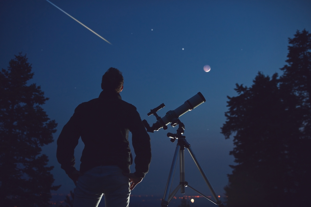 A stargazer standing next to a telescope and looking up at the sky as a meteor shoots overhead
