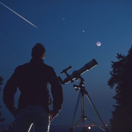 A stargazer standing next to a telescope and looking up at the sky as a meteor shoots overhead